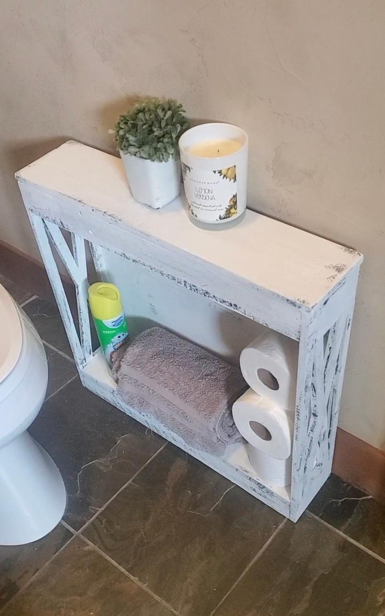 May include: A narrow, white, distressed-finish wooden shelf unit with X-shaped side supports. The top surface holds a small potted green plant and a Lemon Verbena candle. The lower shelf contains a folded gray towel, two toilet paper rolls, and a yellow spray bottle.