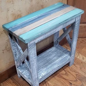 May include: A rectangular wooden side table with a distressed gray finish on the frame and legs. The tabletop features horizontal planks painted in alternating shades of teal, blue, and natural wood. The base includes an X-shaped side support.