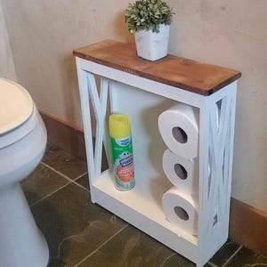 May include: A slim, white wooden bathroom storage shelf with an X-pattern on the sides and a dark brown wooden top. The shelf holds three white toilet paper rolls and a yellow and green aerosol cleaning spray, with a small potted plant on top.