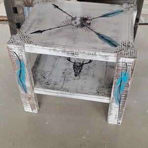 May include: A rustic, distressed white side table with a gray top. The top features crossed arrows and teal feather accents. The lower shelf displays a black deer skull. Teal feather accents are painted on the legs.