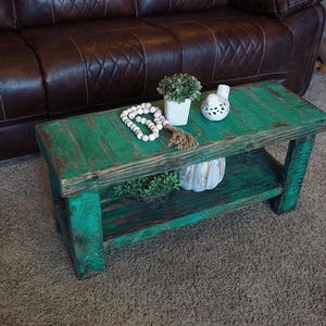 May include: A rustic, green-painted wooden coffee table with a lower shelf. The table is adorned with decorative items, including a small potted plant, a string of white beads, and a decorative bird figurine. The table is approximately 100 cm long and 50 cm wide.