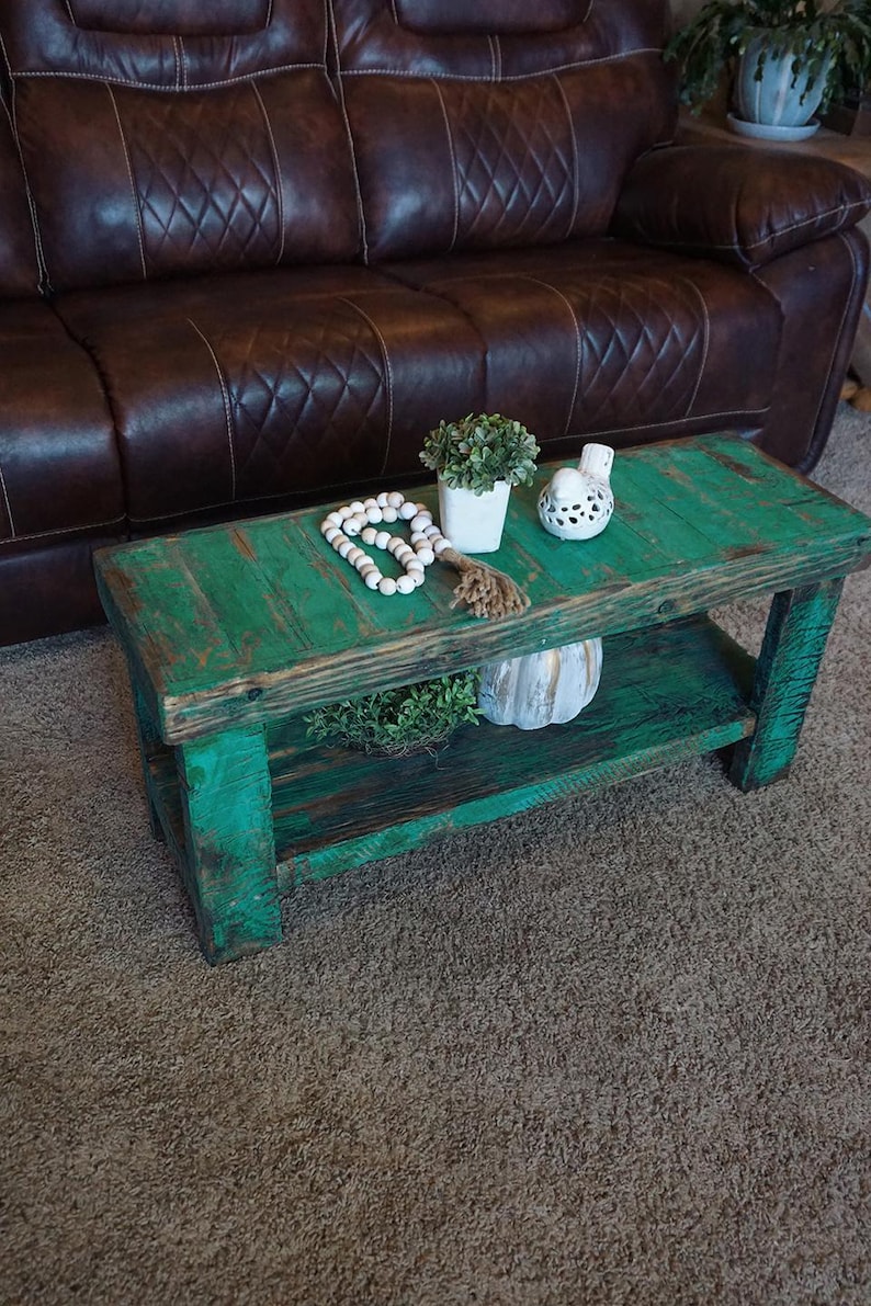 May include: A rustic, green-painted wooden coffee table with a lower shelf. The table is adorned with decorative items, including a small potted plant, a string of white beads, and a decorative bird figurine. The table is approximately 100 cm long and 50 cm wide.