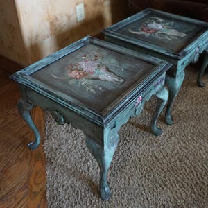 May include: Two square side tables with a distressed teal finish and cabriole legs. The tabletop surfaces feature a painted design of a bull skull adorned with pink and white flowers, set against a dark, weathered background with visible wood grain.