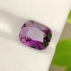 May include: A cushion-cut gemstone with a vibrant purple hue and multifaceted surface. The stone displays deep violet tones and light-reflecting facets throughout its structure, showcasing a clear, transparent quality and a polished finish.