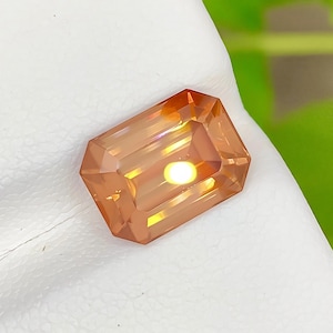 May include: A rectangular, emerald-cut gemstone with a vibrant orange and golden-yellow hue. The stone features precise, multi-faceted edges that reflect light, creating a bright, glowing center point within its translucent, polished surface.