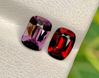 May include: Two faceted gemstones. One is a rectangular cut, deep purple color. The other is a rectangular cut, deep red color. Both are set against a white textured background.