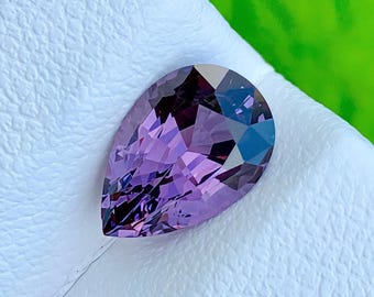 Può includere: Una gemma sfaccettata a forma di pera con una vibrante tonalità viola. La pietra presenta una complessa disposizione di sfaccettature triangolari e quadrangolari che creano riflessi viola intenso e indaco scuro sulla superficie lucida.