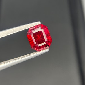 May include: A faceted, rectangular-cut, deep red gemstone held by silver tweezers. The gem has a brilliant, internal starburst pattern. The background is a blurred gradient of gray and black.