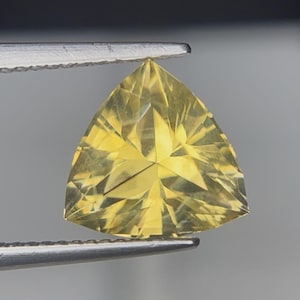 Puede incluir: Una gema amarilla en forma de triángulo, sujeta con pinzas. La gema tiene múltiples facetas que reflejan la luz, creando un efecto brillante. La forma es un triángulo equilátero. Posiblemente una piedra tallada.