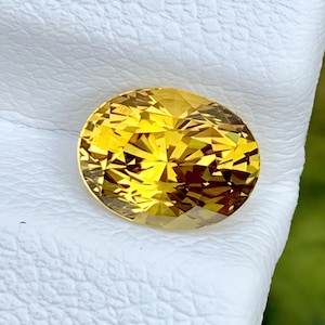 Könnte beinhalten: Ein leuchtend gelber Edelstein in ovaler Form mit Facettenschliff. Der Stein zeigt eine brillante, reflektierende Oberfläche mit zahlreichen geometrischen Facetten, die das Licht einfangen und ein helles, goldenes Erscheinungsbild erzeugen.