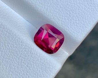 May include: A cushion-cut, faceted gemstone in shades of red and pink. The gem is set against a white, textured surface. The gemstone's facets reflect light, creating a sparkling effect. This item is suitable for jewelry making.