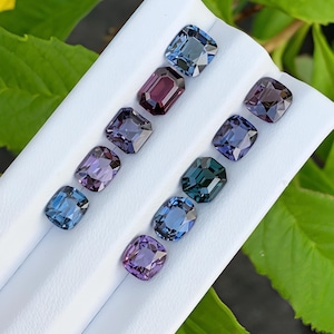 Puede incluir: Una colección de diez piedras preciosas facetadas dispuestas en dos filas sobre un expositor blanco. Las piedras presentan varios cortes, incluyendo formas de cojín y esmeralda, mostrando un espectro de colores que va desde el azul profundo y el morado hasta el verde azulado.