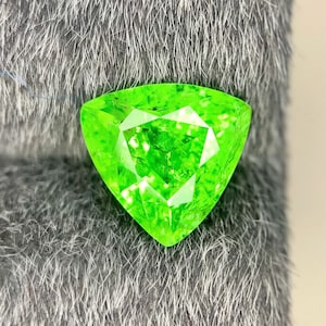 Puede incluir: Una gema triangular de color verde neón brillante. La gema presenta múltiples facetas que reflejan la luz, creando un efecto brillante. La gema se encuentra sobre un fondo gris texturizado.