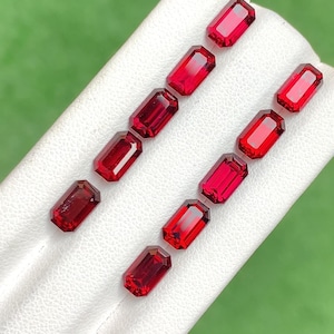 May include: A set of nine red gemstones, cut in a rectangular shape, arranged in two rows on a white surface. The gemstones are all the same size and shape, but they vary in shade of red.