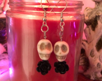 May include: A pair of silver skull earrings with black rose accents. The earrings are hanging from a glass jar with a red candle inside.