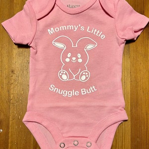 May include: Pink baby onesie with the text "Mommy's Little Snuggle Butt" and a white bunny graphic. The onesie has short sleeves and three snap closures at the bottom. The brand "Hanes" is printed on the collar.