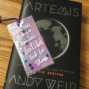 May include: A book titled "Artemis" by Andy Weir, a New York Times Bestselling author of "The Martian". The book cover features a black and white image of the moon. A bookmark with a purple and blue background and glitter accents reads "When it rains look for rainbows When it's dark look for stars".