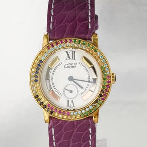 May include: A gold-tone wristwatch with a white dial featuring the text 'must de Cartier', Roman numerals at twelve and six, and a bezel set with multi-colored gemstones. The watch has a purple textured leather strap with white stitching.