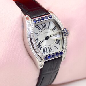 May include: A silver Cartier watch with a white face and black leather band. The watch has a diamond bezel and blue sapphire accents. The watch face has Roman numeral hour markers and the words 'CARTIER' printed on it.
