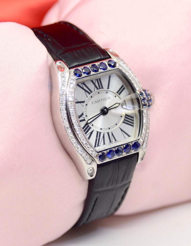 May include: A silver Cartier watch with a white face and black leather band. The watch has a diamond bezel and blue sapphire accents. The watch face has Roman numeral hour markers and the words 'CARTIER' printed on it.