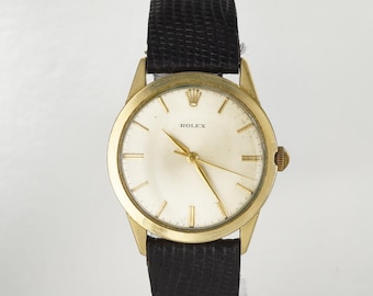 May include: A gold-tone wristwatch featuring a round cream-colored dial with gold-tone hour markers and hands. The watch face displays the Rolex logo and name. It is attached to a black textured leather strap with a visible grain pattern.