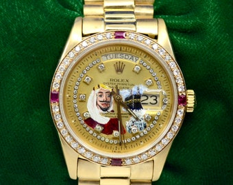 Puede incluir: Un reloj Rolex Day-Date de oro con un bisel engastado con diamantes y una esfera pintada con un hombre con una corona. La esfera del reloj muestra el día "TUESDAY" y la fecha "23".