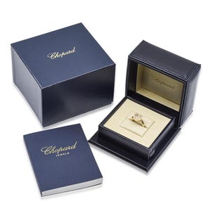 May include: A gold ring with a heart-shaped diamond is displayed in a black leather jewelry box with a cream-colored interior. The box is open, revealing the ring. A navy blue box and a navy blue booklet with the word "Chopard" in gold lettering are also present.