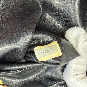 May include: Close-up of a black leather handbag interior. A small, rectangular tag with the number "0649475" is visible. The leather has a textured, pebbled appearance. The bag's zipper and a portion of a gold-colored clasp are also visible.