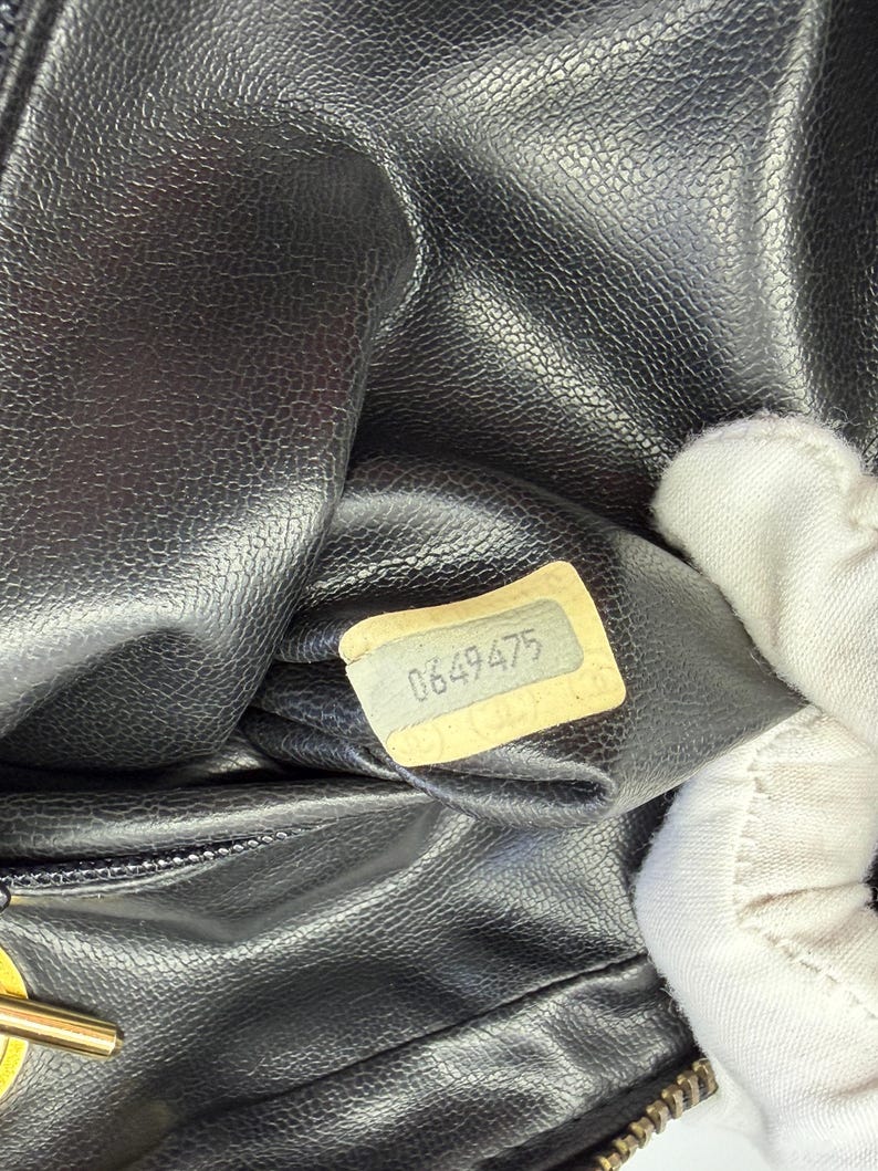 May include: Close-up of a black leather handbag interior. A small, rectangular tag with the number "0649475" is visible. The leather has a textured, pebbled appearance. The bag's zipper and a portion of a gold-colored clasp are also visible.