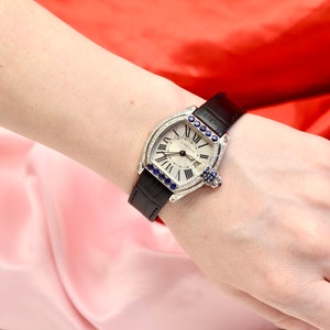 May include: A silver watch with a white face and black leather band. The watch is decorated with diamonds and blue sapphires around the bezel. The watch face has Roman numeral hour markers and a date window at the 3 o'clock position. The watch is a Cartier model.