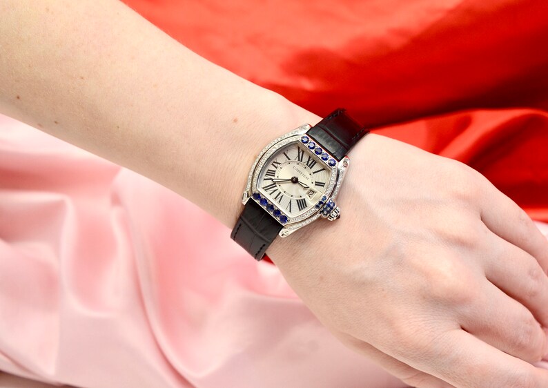 May include: A silver watch with a white face and black leather band. The watch is decorated with diamonds and blue sapphires around the bezel. The watch face has Roman numeral hour markers and a date window at the 3 o'clock position. The watch is a Cartier model.