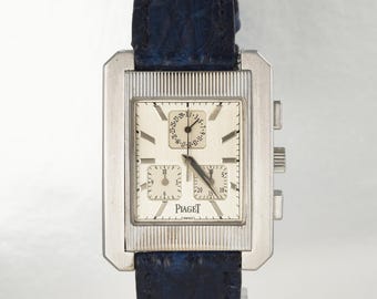 May include: A rectangular silver-tone wristwatch with a cream-colored dial, featuring three sub-dials, silver baton hour markers, and the brand name Piaget printed in black. The watch is attached to a dark blue textured leather strap.