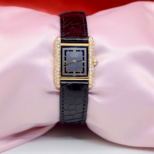 May include: A gold Cartier watch with a black face and diamond encrusted bezel. The watch is on a black leather band.