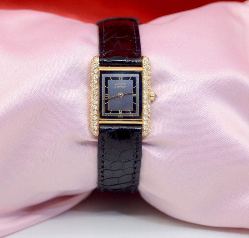 May include: A gold Cartier watch with a black face and diamond encrusted bezel. The watch is on a black leather band.