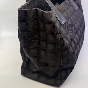 May include: Black tote bag with a checkered pattern and leather handles. The bag features a repeating logo design. The bag is made of a soft, flexible material and has a rectangular shape.