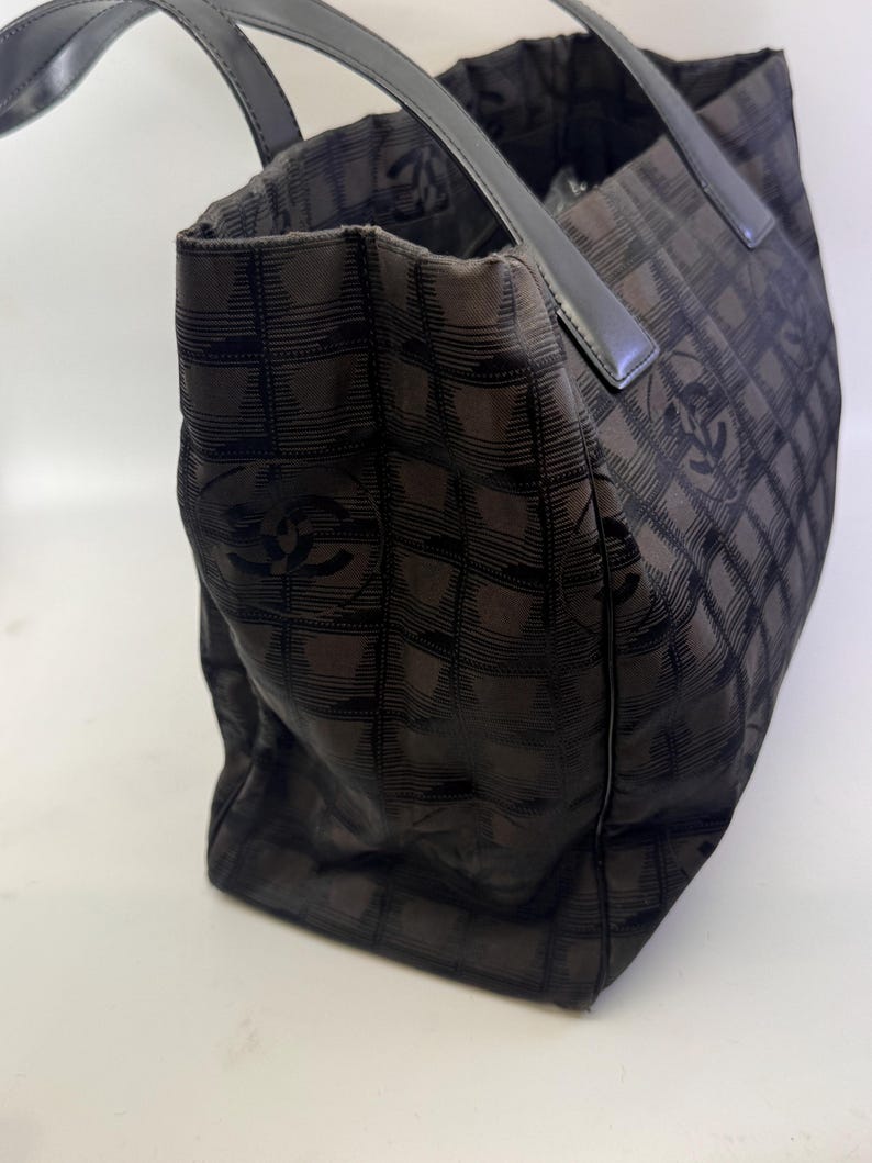 May include: Black tote bag with a checkered pattern and leather handles. The bag features a repeating logo design. The bag is made of a soft, flexible material and has a rectangular shape.