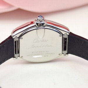 May include: A silver Cartier Roadster watch with a black leather band. The back of the watch has the words 'Cartier Roadster' engraved on it, along with the words 'Stainless Steel' and 'Water Resistant 30M'.
