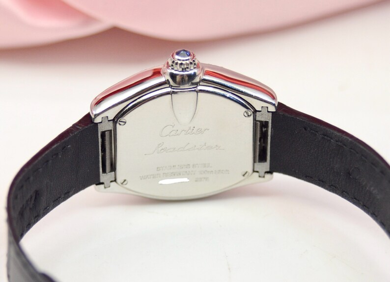 May include: A silver Cartier Roadster watch with a black leather band. The back of the watch has the words 'Cartier Roadster' engraved on it, along with the words 'Stainless Steel' and 'Water Resistant 30M'.