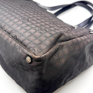 K&ouml;nnte beinhalten: Eine schwarze und braune Tragetasche mit sich wiederholendem Muster. Die Tasche hat schwarze Griffe und ein silbernes KnopfdDetail. Der Stoff der Tasche hat ein gitterartiges Muster mit einem Logo-Design.