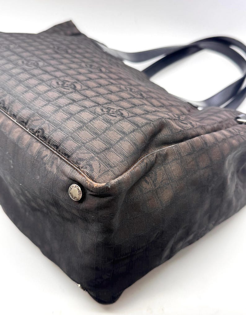 K&ouml;nnte beinhalten: Eine schwarze und braune Tragetasche mit sich wiederholendem Muster. Die Tasche hat schwarze Griffe und ein silbernes KnopfdDetail. Der Stoff der Tasche hat ein gitterartiges Muster mit einem Logo-Design.