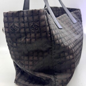 K&ouml;nnte beinhalten: Braune Tragetasche mit sich wiederholendem quadratischem Muster und Logo-Design. Die Tasche hat schwarze Ledergriffe und einen silberfarbenen Metallring. Sie besteht aus einem strukturierten Stoff.