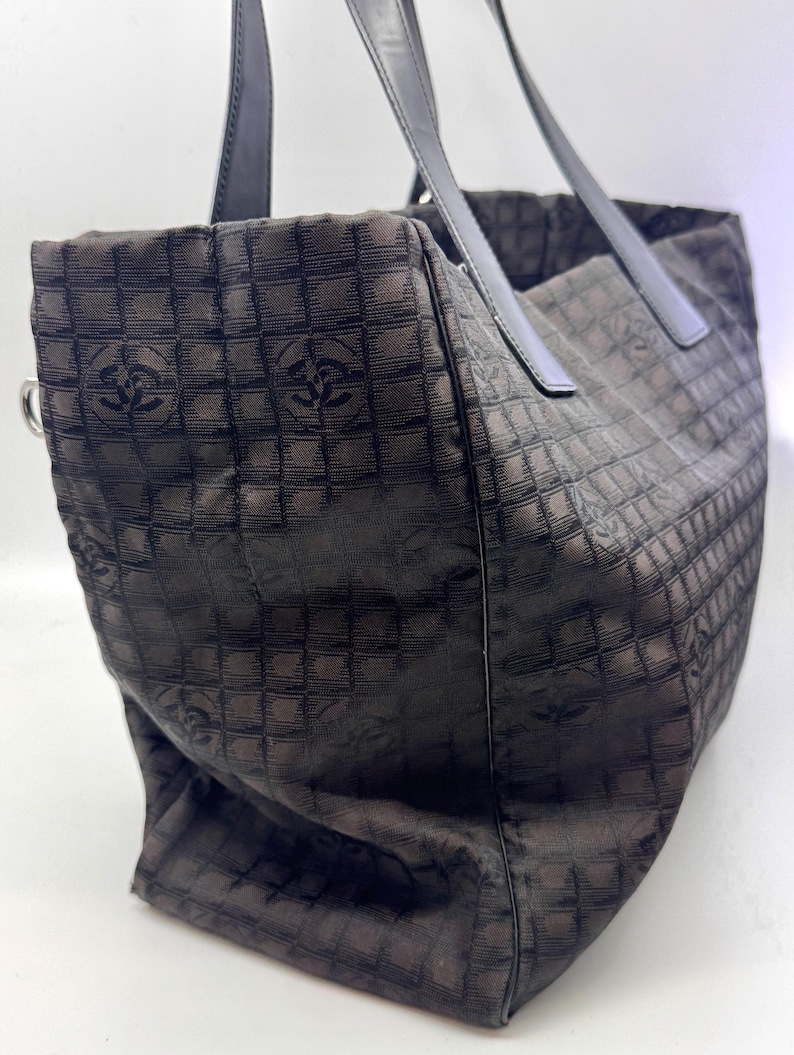 K&ouml;nnte beinhalten: Braune Tragetasche mit sich wiederholendem quadratischem Muster und Logo-Design. Die Tasche hat schwarze Ledergriffe und einen silberfarbenen Metallring. Sie besteht aus einem strukturierten Stoff.