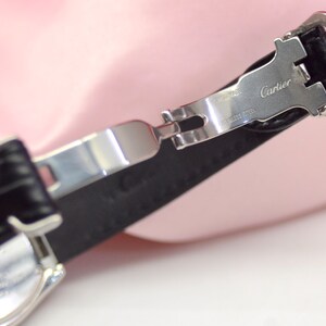 May include: A silver watch band with a black leather strap. The band has a clasp that says 'Cartier' and 'Stainless Steel'.