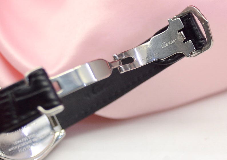 May include: A silver watch band with a black leather strap. The band has a clasp that says 'Cartier' and 'Stainless Steel'.