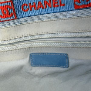 Puede incluir: Interior de bolso azul claro con una correa con el logo azul y rojo que dice "CHANEL". El interior presenta un forro azul claro, una cremallera y una etiqueta de cuero azul.