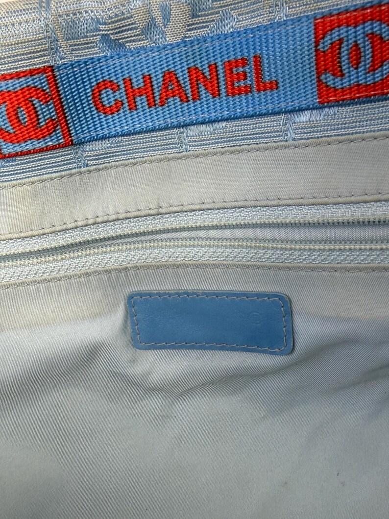 Puede incluir: Interior de bolso azul claro con una correa con el logo azul y rojo que dice "CHANEL". El interior presenta un forro azul claro, una cremallera y una etiqueta de cuero azul.