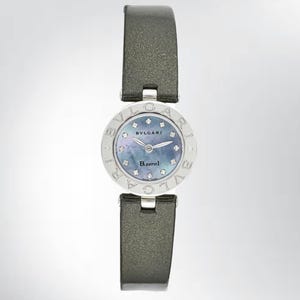 May include: A silver-toned Bvlgari B.zero1 watch with a gray leather strap. The watch face has a blue mother-of-pearl dial with diamond accents and the brand name. The watch is a luxury item.