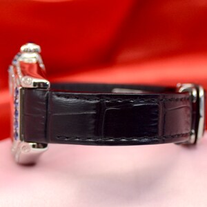 May include: A silver wristwatch with a black leather band. The watch face is surrounded by small blue gemstones.