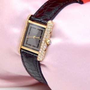 May include: A gold Cartier watch with a black leather band and a diamond-encrusted bezel. The watch face is black with Roman numeral hour markers and the word 'Cartier' printed in white.