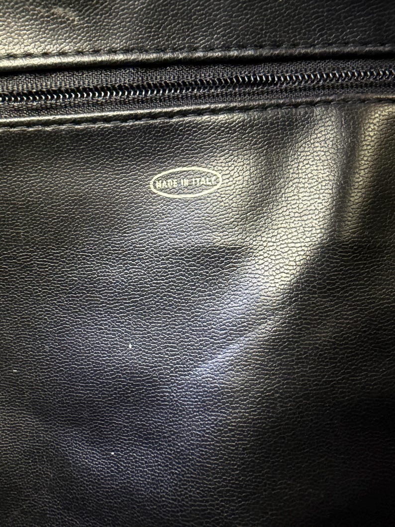 May include: Close-up of a black leather item with a zipper and the text "MADE IN ITALY" in a white oval. The leather has a textured surface.